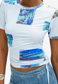Top corto de manga corta en azul y blanco que presenta patrones abstractos en tonos de azul, verde y negro; tejido texturizado con fruncidos en los laterales.