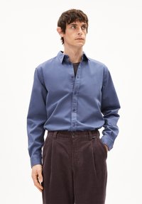 Camicia blu con bottoni in cotone, dotata di colletto e maniche lunghe. Abbinata a pantaloni in velluto a coste marroni, che mostrano righe verticali e pieghe.