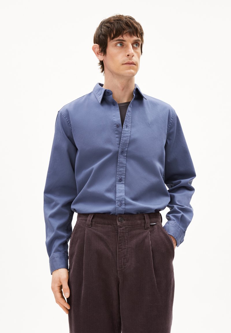 Camicia blu con bottoni in cotone, dotata di colletto e maniche lunghe. Abbinata a pantaloni in velluto a coste marroni, che mostrano righe verticali e pieghe.