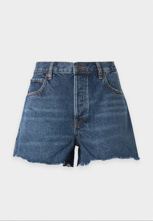 Tânără stă în picioare purtând o bluză albă scurtă cu nasturi și pantaloni scurți din denim albastru cu margini destrămate, cu o mână în buzunar.