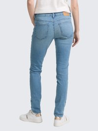 Oseba oblečena v svetloblue skinny jeans in bele superge z metalnimi akcenti, ki stoji ob enostavni svetli ozadju.