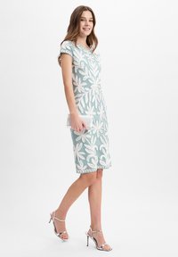 Helles blaues, tailliertes Kleid mit weißer Blumenstickerei, kurzen Ärmeln und gewelltem Saum. Kombiniert mit silbernen High-Heelsandalen und einer Clutch.