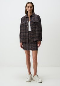 Femme portant un blazer à carreaux texturé sombre et une jupe assortie avec des baskets blanches, se tenant devant un fond clair uni.