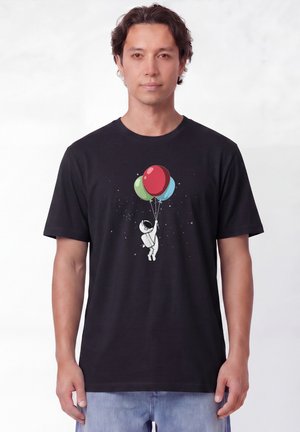 Homme portant un t-shirt noir avec un dessin d'un petit astronaute tenant des ballons rouges, verts et bleus flottant dans l'espace.