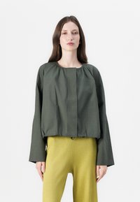 Veste courte vert olive avec un col rond, des détails froncés et des manches longues, associée à un pantalon jaune texturé à côtes.