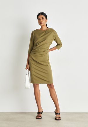 Olive green dress with a boat neckline, three-quarter sleeves, and gathered detail. Paired with black strappy heels and a white handbag.