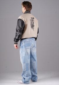 Beige und schwarze Collegejacke mit besticktem Design auf der Rückseite, kombiniert mit hellblauen Patchwork-Jeans und weißen Sneakers.