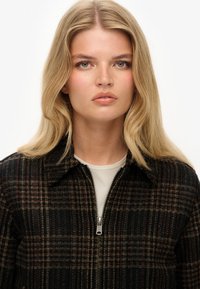 Ruitjes zip-up jack in donkere tinten, met een kraag, gestructureerde stof en zilveren ritsdetails. Onderlaag is een neutraal top.