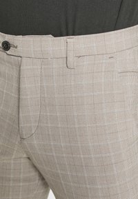 Beige rutiga byxor med en strukturerad finish, med knappstängning, bältesslingor och sidofickor, som framhäver ett subtilt rutig mönster.