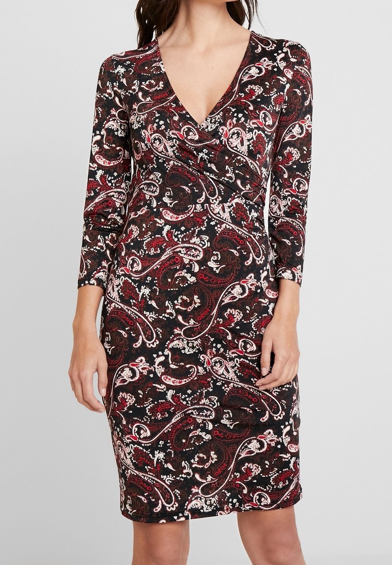 Kvinna som bär en figursydd klänning i knälängd med långa ärmar och ett röd, svart och vitt paisleymönster, med en V-ringning.