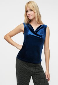 Mørkeblå ermeløs topp i myk, glanset stoff med drapert hals; kombinert med teksturerte mønstrede sorte bukser.