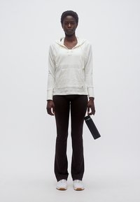 Γυναίκα που στέκεται με το σώμα προς τα εμπρός, φορώντας λευκή φούτερ με κουκούλα, μαύρο παντελόνι, λευκά αθλητικά παπούτσια, κρατώντας ένα μαύρο μπουκάλι νερού στο δεξί της χέρι.