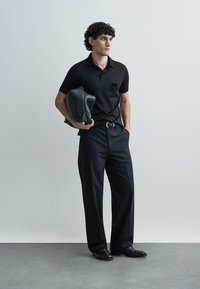 Black polo shirt with emblem, black wide-leg trousers, and black shoes. Model holding a dark grey jacket, standing against a neutral background.