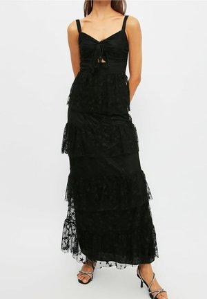 Kobieta ubrana w czarną, warstwową maxi sukienkę z koronki z cienkimi ramiączkami, wiązaniem z przodu i wzorami gwiazd, w parze z czarnymi sandałami na wysokim obcasie z paskami.