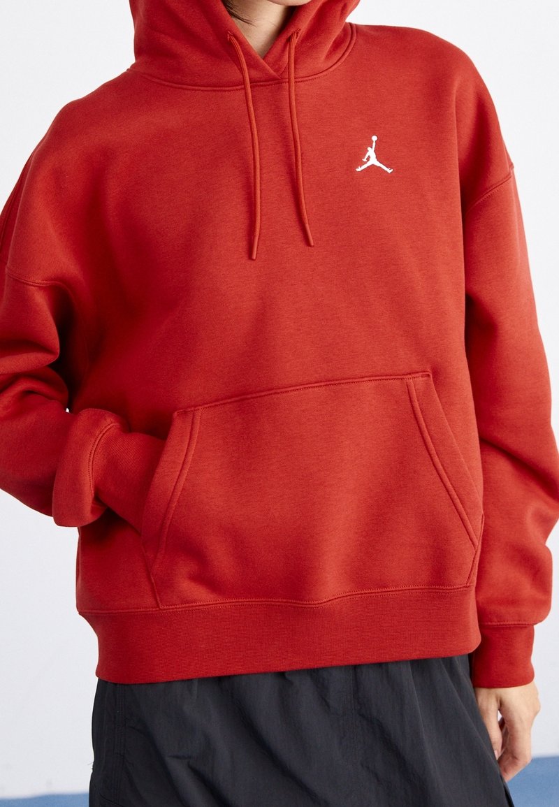 Personne portant un sweat à capuche rouge avec un logo Jumpman blanc et un pantalon noir, les mains dans les poches du sweat, sur un fond uni.