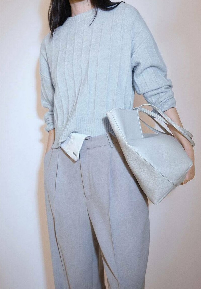 Personne portant un pull côtelé gris clair et un pantalon taille haute assorti, tenant un sac à main structuré gris clair par les anses.