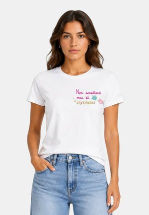 Donna con capelli castani che indossa una T-shirt bianca con testo italiano e piccole illustrazioni colorate di borse, abbinata a jeans blu.