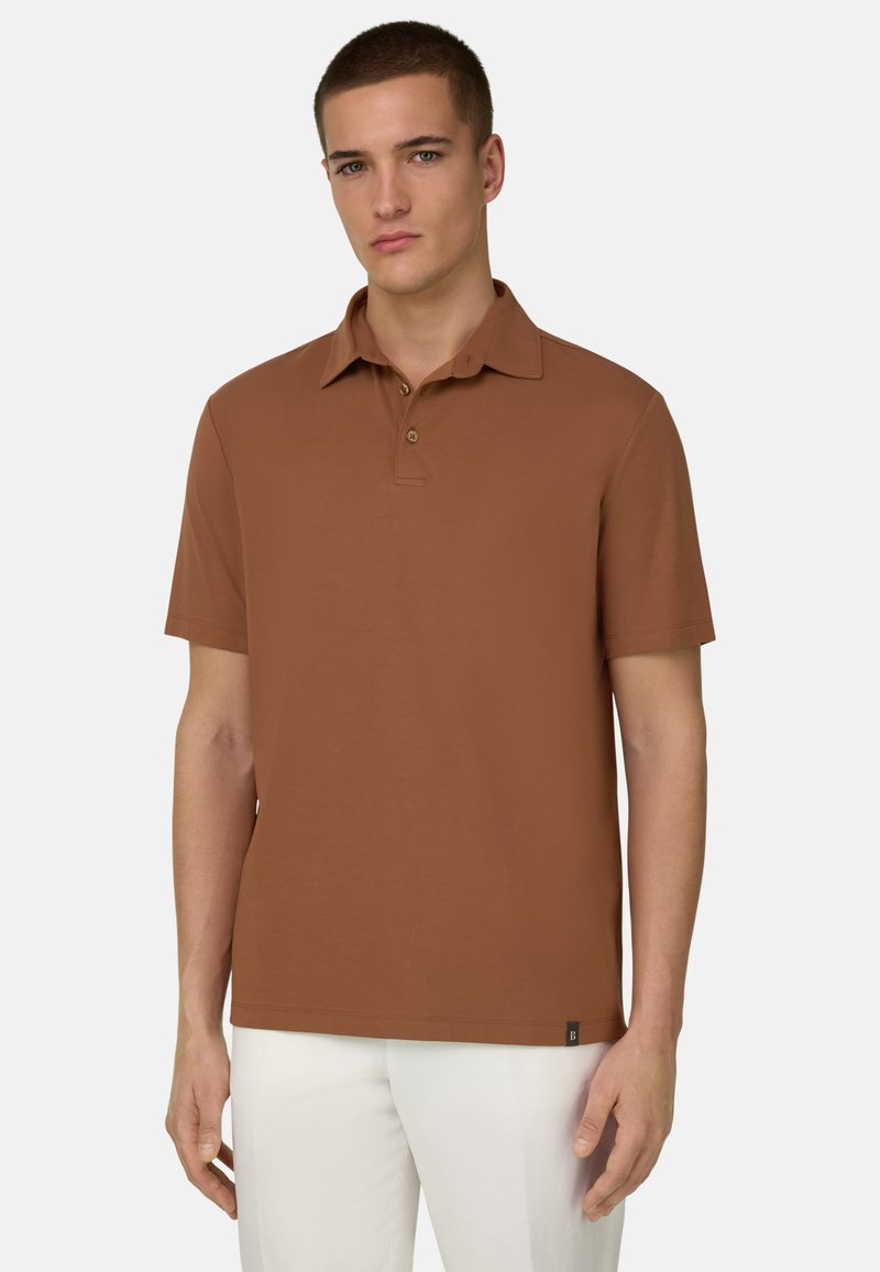 Jeune homme portant un polo marron à manches courtes et un pantalon blanc, sur un fond clair uni.