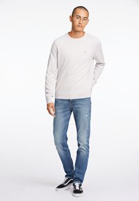 Lichtgrijze crewneck trui met geribde mouwen, gecombineerd met verwaarloosde blauwe jeans en zwart-witte sneakers. Minimale logo-detail op de trui.