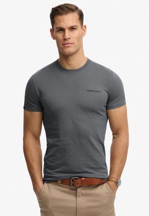 Mężczyzna ubrany w dopasowaną ciemnoszarą koszulkę Superdry & Co, beżowe spodnie z brązowym skórzanym paskiem oraz zegarek na nadgarstku, stojący z rękami w kieszeniach.