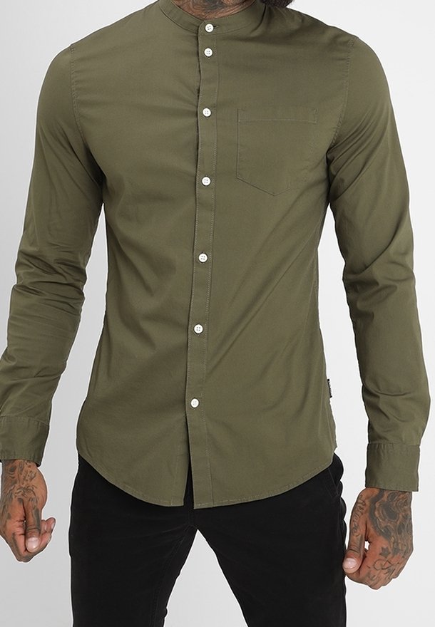 Homme portant une chemise ajustée vert olive à col mandarin et un pantalon noir, montrant des mains et des bras tatoués.