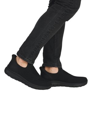 Zapatillas de malla negra sin cordones con diseño texturizado, suela flexible y talón acolchado. Llevadas con jeans oscuros, resaltando el contorno del zapato.