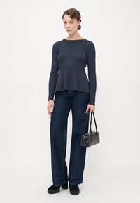 Kvinne iført mørk blå langermet topp og vide jeans, holder en liten svart veske, stående mot en enkel hvit bakgrunn.