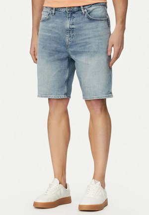 Uomo che indossa pantaloncini di jeans azzurri abbinati a scarpe da ginnastica bianche basse, su sfondo semplice.