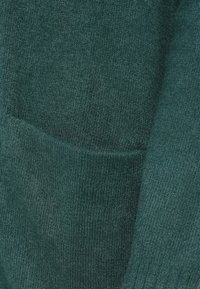Maglione verde lavorato a maglia con una consistenza morbida, dotato di una tasca frontale e polsini a costine. Il primo piano rivela dettagli di cucitura fini.