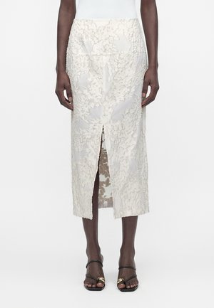 Άτομο που φοράει λευκή midi φούστα από δαντέλα με λουλούδια και κεντρική σχισμή μπροστά, καθώς και μαύρα σανδάλια με λουράκια και χρυσές λεπτομέρειες.