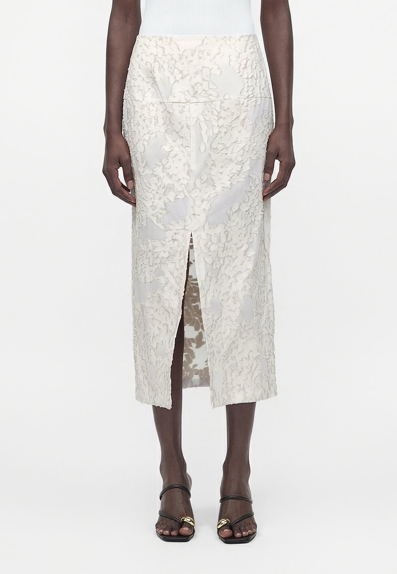 Άτομο που φοράει λευκή midi φούστα από δαντέλα με λουλούδια και κεντρική σχισμή μπροστά, καθώς και μαύρα σανδάλια με λουράκια και χρυσές λεπτομέρειες.