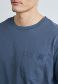 Heren T-shirt met korte mouwen in leisteenkleurige katoen. Voorzien van een borstzak met een klein CK-logo. Klassieke ronde halslijn en een zachte textuur.