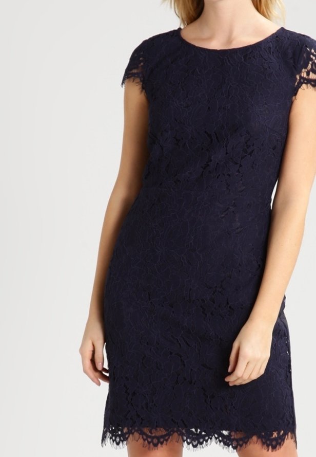 Femme portant une robe en dentelle bleu marine ajustée avec des manches courtes et un ourlet festonné, debout devant un fond uni.