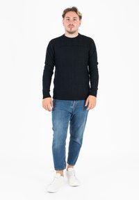 Czarny sweter z teksturowanym splotem, z okrągłym dekoltem, zestawiony z niebieskimi dżinsami o slim-fit i białymi trampkami. Styl jest casualowy i nowoczesny.