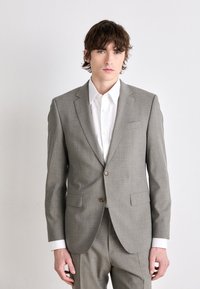 Abito grigio chiaro con un motivo testurizzato, giacca monopetto, revers a punta e dettagli con bottoni marroni. Camicia bianca visibile sotto.