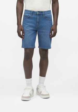 Mann iført blå denimshorts, hvite ankelsokker, hvite og beige joggesko, stående foran hvit ensfarget bakgrunn.