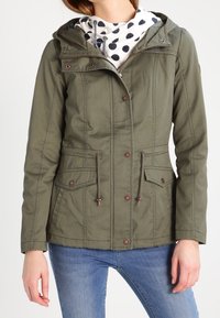 Oliwkowa zielona parka z kapturem, zapinana na zamek i zatrzaski, z paskiem ściągającym w talii, dwoma przednimi kieszeniami i teksturowaną tkaniną. Biały szalik w grochy.