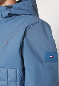 Modrá bunda s kapucí, s malým vlajkovým logem na prsou a obdélníkovou nášivkou značky na horním rukávu.