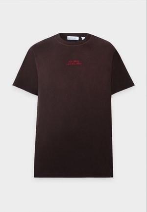 Uomo in piedi che indossa una maglietta marrone scuro, pantaloni neri con una grafica sulla coscia e scarpe da ginnastica bianche e nere su uno sfondo semplice.