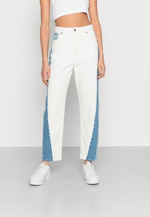 Femme portant un jean blanc taille haute avec des panneaux latéraux bleus, assorti à des baskets blanches à plateforme et un haut court blanc.