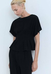 Černý peplum top vyrobený z texturované tkaniny, s krátkými rukávy, jemnými detailem šití a uvolněným střihem. V kombinaci s černými kalhotami.
