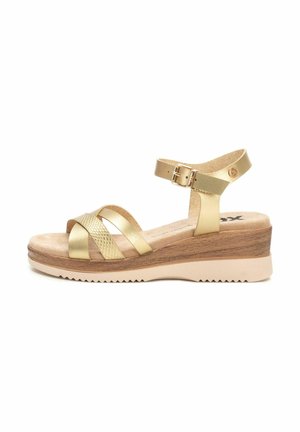Chanclas doradas con correas cruzadas texturizadas, un cierre en el tobillo y una suela de cuña con acabado similar a la madera y base de goma beige.