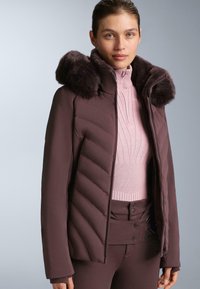 Smeđa protkana jakna s kapuljačom od lažnog krzna, uparena s lagano ružičastim rebrastim džemperom i pripadajućim visokim hlačama. Glatka tekstura, uski kroj.