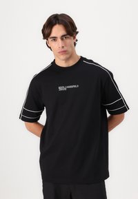 Črn bombažni t-shirt z okroglim izrezom, belim logotipom na prsih in belimi oblogami ob rokavih. Ravan kroj s kratkimi rokavi.