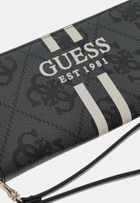 Portafoglio nero con motivo, realizzato in materiale testurizzato. Presenta strisce bianche e il logo "GUESS EST 1981". Include una tracolla staccabile.