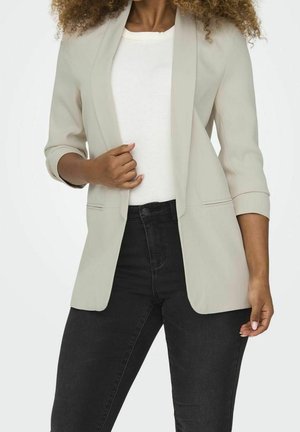 Vrouw met lichtbeige blazer met opgerolde mouwen, wit shirt en zwarte hoge taille jeans tegen een effen achtergrond.