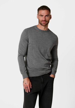 Man met kort haar en baard, die een grijze trui met lange mouwen en een zwarte broek draagt, staat met één hand in zijn zak tegen een witte achtergrond.