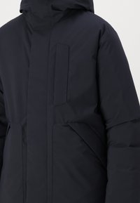 Mørkeblå puffermantel med høj krave, store forlommer, en glat tekstur og et elegant, minimalistisk design.