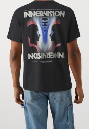 Personne portant un t-shirt noir avec des silhouettes en miroir qui se font face, dégradé bleu et rouge, et texte en gras « INNERVISION » au dos.