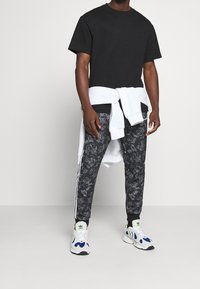 Man som bär en svart t-shirt, svarta mönstrade joggers med vita ränder, vita sneakers och en vit sweatshirt knuten runt midjan.